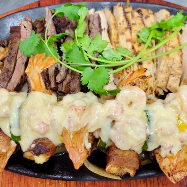 Sizzling platter of grilled steak, chicken, and bacon-wrapped shrimp with melted cheese and cilantro.