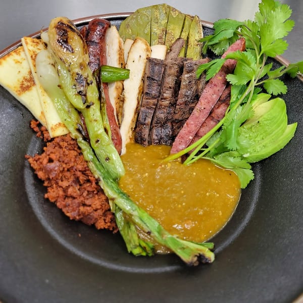Mexican dish with grilled meats, chorizo, cheese, nopales, avocado, and salsa verde in a black bowl.